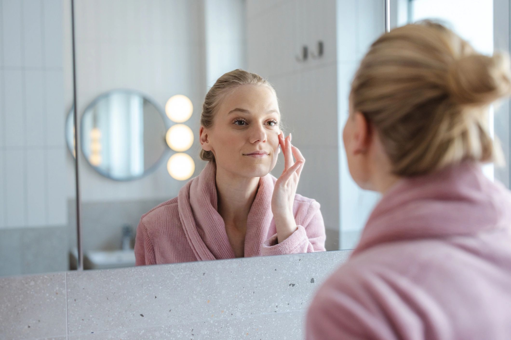 A woman in a pink robe gently applies facial cream while looking at her reflection in a bathroom mirror
