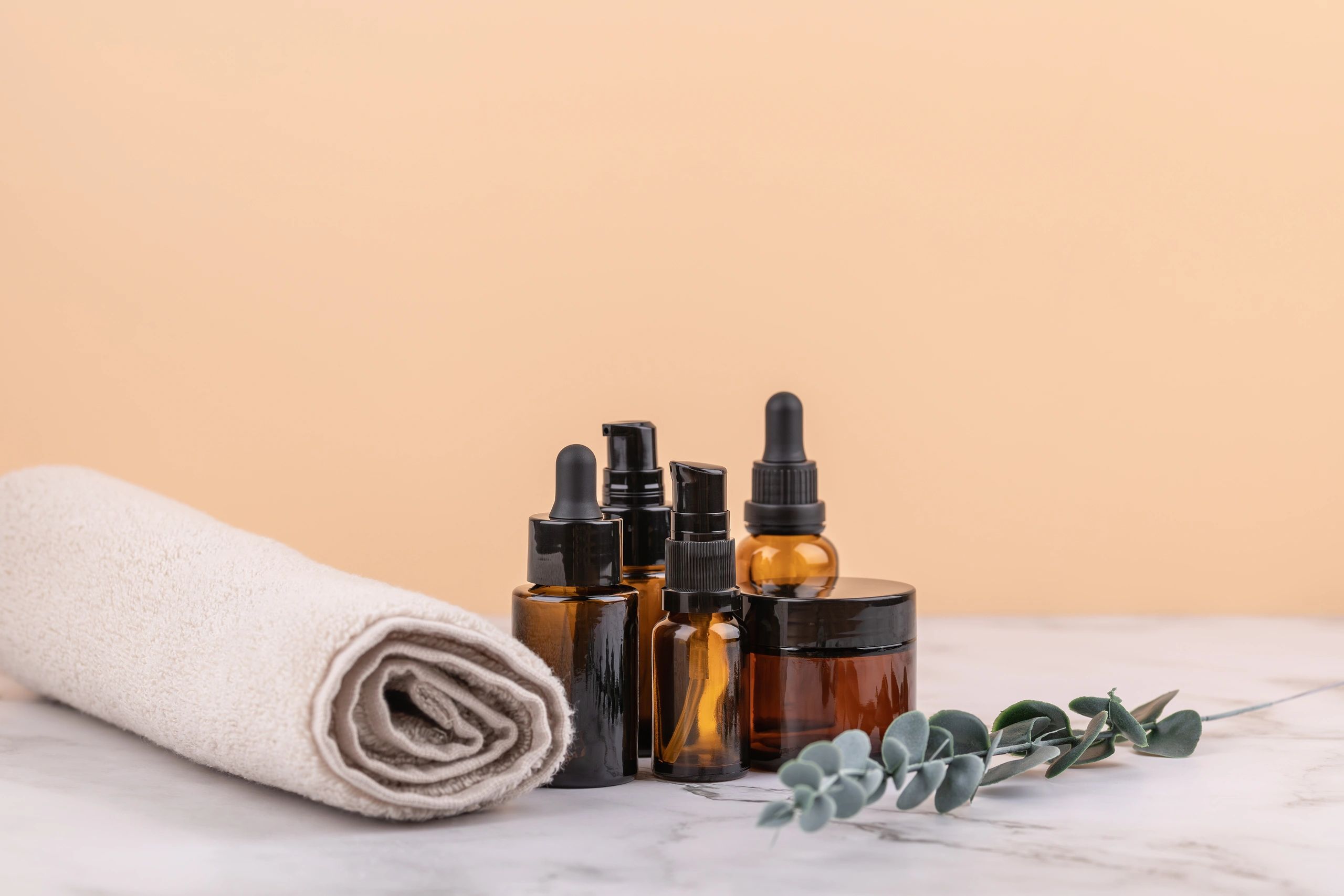 Amber glass bottles with dropper and cosmetic jar beside rolled towel and eucalyptus branch on marble surface